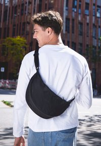 Ung man i vit långärmad skjorta och blå jeans som går utomhus med en svart axelväska över ryggen, urbana byggnader i bakgrunden.