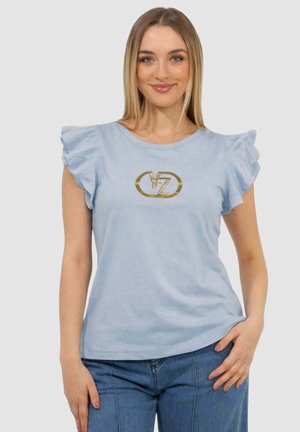 Giovane donna con indosso una maglia azzurra a maniche arricciate con logo dorato "Y/Z", abbinata a jeans blu, in piedi davanti a uno sfondo semplice.