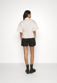 Blusa de manga curta, com padrão floral, em creme, com cintura franzida. Combinada com calções pretos e botas de tornozelo pretas. Fundo branco simples.