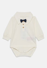 Cremefarbenes Baby-Overall mit langen Ärmeln, einem Kragen, drei Holzknöpfen und einer kleinen dunkelblauen Fliege am Ausschnitt.