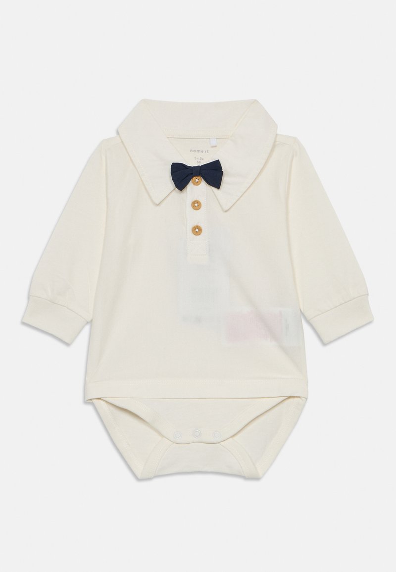 Cremefarbenes Baby-Overall mit langen Ärmeln, einem Kragen, drei Holzknöpfen und einer kleinen dunkelblauen Fliege am Ausschnitt.