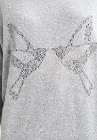 Pull gris clair à texture lisse, orné d'un papillon décoratif en strass sur le devant.