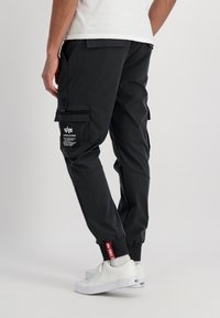 Man med svarta cargojoggingbyxor med märkta fickor och röda lappar, kombinerat med vita lågt skurna sneakers och en vit t-shirt.