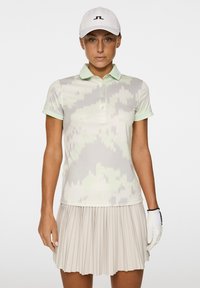 Koszulka polo z krótkim rękawem w jasnoszarym i miętowym kolorze z geometrycznym wzorem, w zestawie z plisowaną beżową spódnicą. Biała czapka z logo.