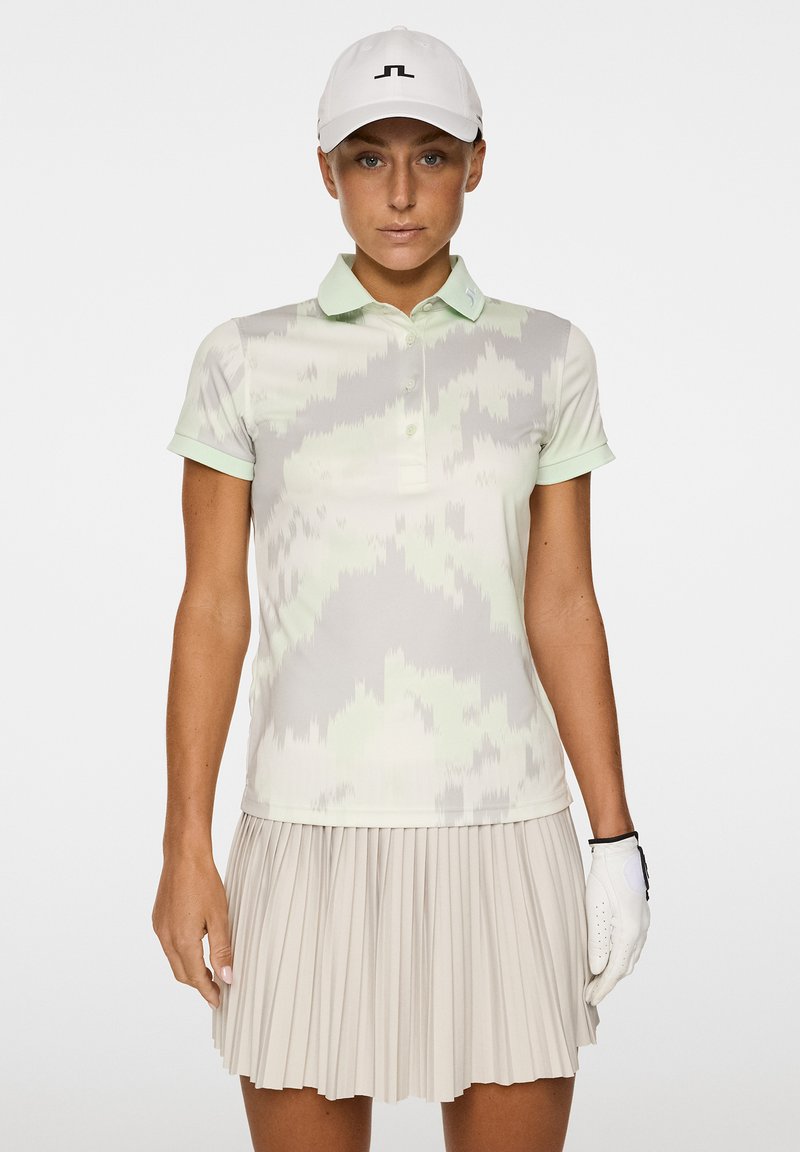 Koszulka polo z krótkim rękawem w jasnoszarym i miętowym kolorze z geometrycznym wzorem, w zestawie z plisowaną beżową spódnicą. Biała czapka z logo.
