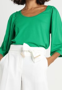 Groene blouse met driekwartmouwen en geplooide details, gecombineerd met witte broeken voorzien van een strik en metalen accenten.