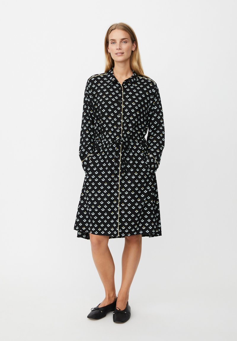 Czarna sukienka z powtarzającym się białym wzorem małych kształtów, długimi rękawami, zapinana na guziki z przodu, z wiązanym w talii paskiem oraz bocznymi kieszeniami.
