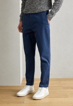 Person, die marineblaue Hose, grauen Strickpullover und weiße Turnschuhe trägt, steht auf einem Holzboden vor einer neutralen Wand.