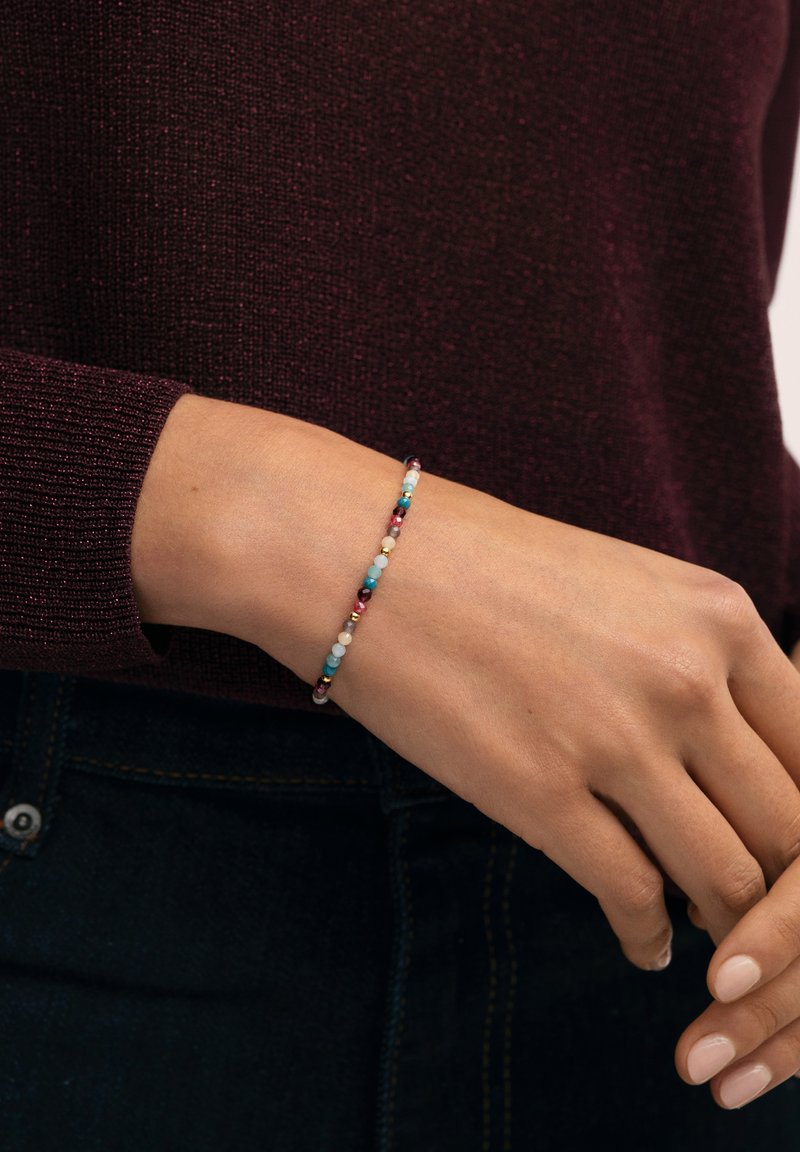 Pulsera de cuentas que presenta una mezcla de cuentas turquesas, rojas, doradas y blancas en un cordón flexible, llevada en una muñeca sobre un suéter oscuro.