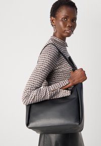 Borsa a spalla nera in pelle con una superficie liscia, forma rettangolare e dettagli di cucitura sottili. Il modello indossa un top con pattern e maniche lunghe.