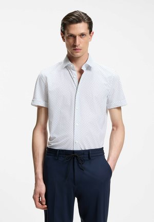 Mand iført hvid kortærmet skjorte med små sorte prikker og marineblå bukser med snøre, stående mod en hvid baggrund.