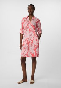 Różowa sukienka z krótkim rękawem w kwiatowy biały wzór, z dekoltem w serek i bocznymi kieszeniami. Modelka w płaskich plecionych sandałach.