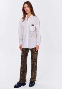 Biała koszula z guzikami i kieszonką na piersi; posiada nadrukowany tekst i grafikę. W połączeniu z rozszerzanymi spodniami w leopardzi wzór i masywnymi czarnymi butami.