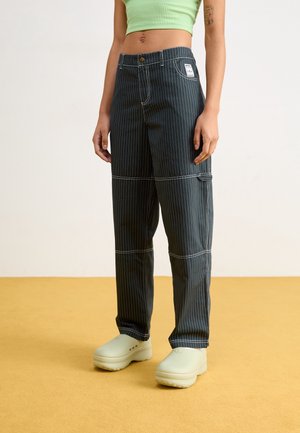 Joven vistiendo pantalones cargo de rayas negras, un top corto verde y zuecos blancos sin cordones, de pie sobre un suelo amarillo.