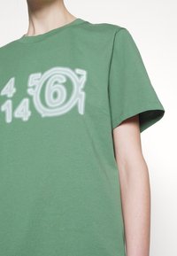 Groene katoenen t-shirt met een grote, gestileerde graphic met cijfers en cirkelvormige vormen in lichtgrijs. Korte mouwen en een ronde hals.