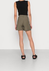 Khaki šortky s přizpůsobeným střihem, vyrobené z lehkého materiálu, s středním páskem a bez viditelného kovového zapínání. Nosené s černým tílkem.