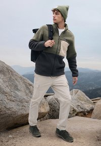 Fleece jacket in olive and black, with a beige patch pocket. Paired with cream pants and dark green shoes; a black backpack and beanie complete the look.