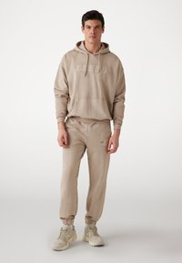 Felpa beige con tasca frontale e logo, abbinata a pantaloni sportivi tapered coordinati. Entrambi realizzati in tessuto morbido, con polsini elasticizzati.