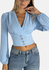 Hellblaues, langärmliges, kurz geschnittenes Oberteil mit tiefem V-Ausschnitt, strukturierter Stoff und vier Holzknöpfen an der Taille. Kombiniert mit hoch taillierten Jeans.