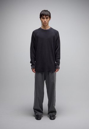 Mladá osoba s krátkymi tmavými vlasmi stojí čelom vpred, oblečená v čiernej dlhorukávovej košeli, voľných sivých nohaviciach a čiernych topánkach.