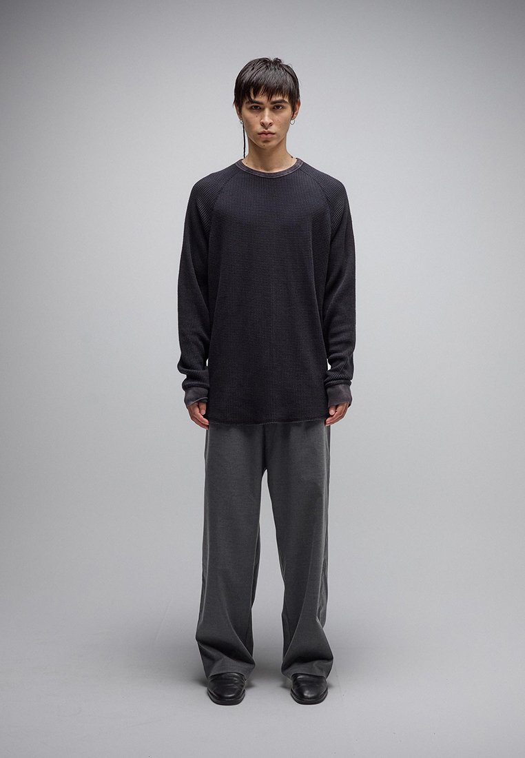 Νεαρό άτομο με κοντά σκούρα μαλλιά στέκεται αντικρίζοντας μπροστά φορώντας μαύρο πουκάμισο μακρυμάνικο, φαρδιά γκρι παντελόνια και μαύρα παπούτσια.