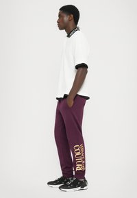 Polo branca de manga curta com colarinho e punhos pretos, emparelhada com calças justas em cor bordô com "VERSACE JEANS COUTURE" em letras douradas. Ténis pretos.