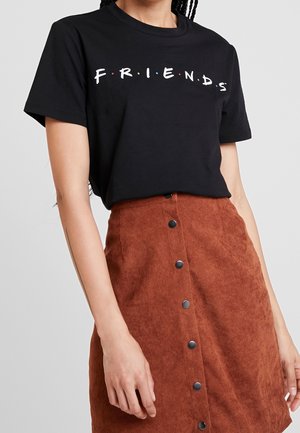 Kobieta ubrana w czarną koszulkę z logo "FRIENDS" wpuszczoną w przypiętą spódnicę z rdzawego sztruksu, stojąca z rozluźnionymi ramionami.