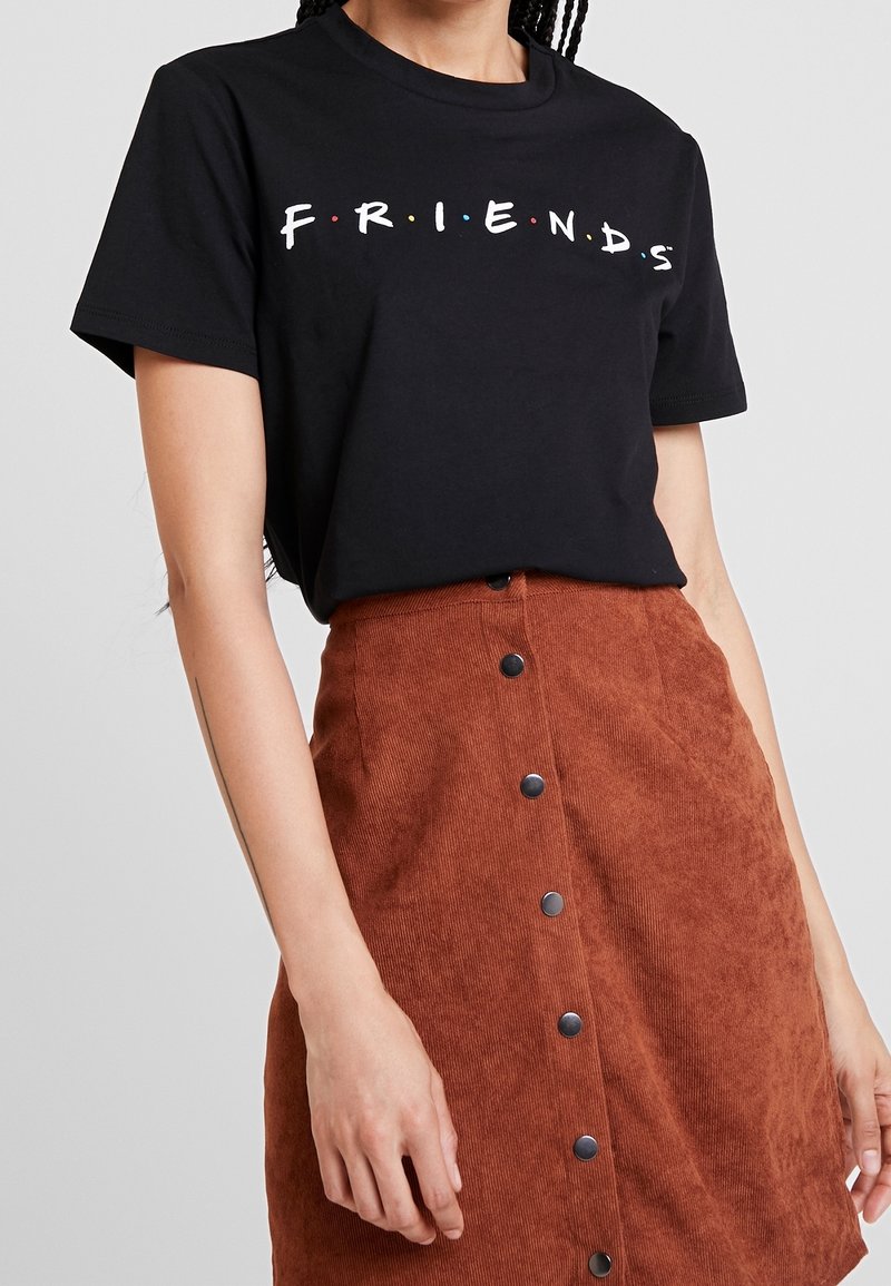 Žena v černém tričku s nápisem "FRIENDS" zastrčeném do zapnuté cihlově zbarvené manšestrové sukně, stojící s uvolněnýma rukama.