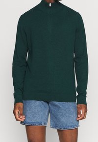 Mörkgrön stickad tröja med kvartdragkedja, långa ärmar och ribbade muddarna, tillsammans med denimshorts. Mjuk textur, åtsittande design.