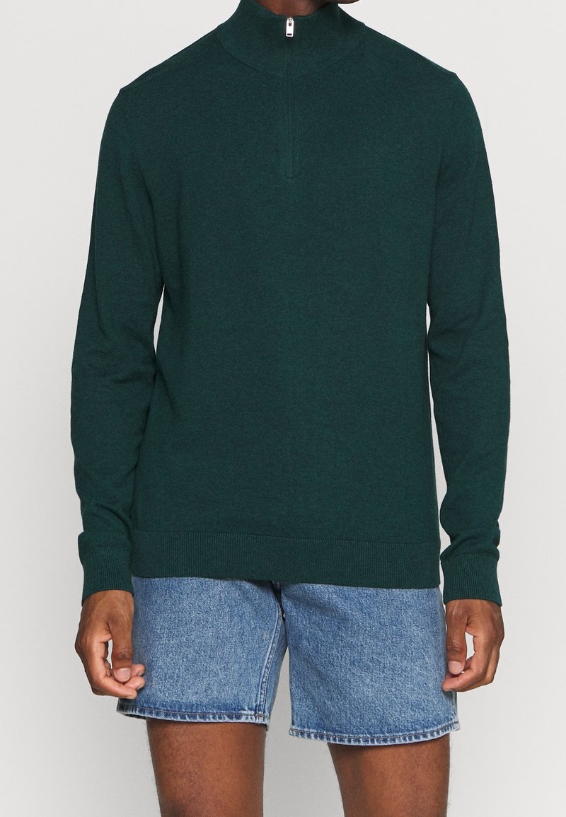 Mörkgrön stickad tröja med kvartdragkedja, långa ärmar och ribbade muddarna, tillsammans med denimshorts. Mjuk textur, åtsittande design.