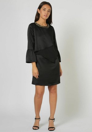 Mujer con vestido de satén negro con escote adornado con cuentas y mangas acampanadas, combinado con sandalias negras de tacón alto con correa en el tobillo, de pie.
