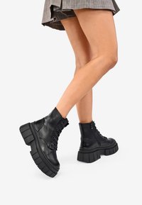 Gambe in stivali neri con piattaforma chunky e lacci, con cerniere, in piedi su uno sfondo bianco, indossando una gonna a quadri corta.