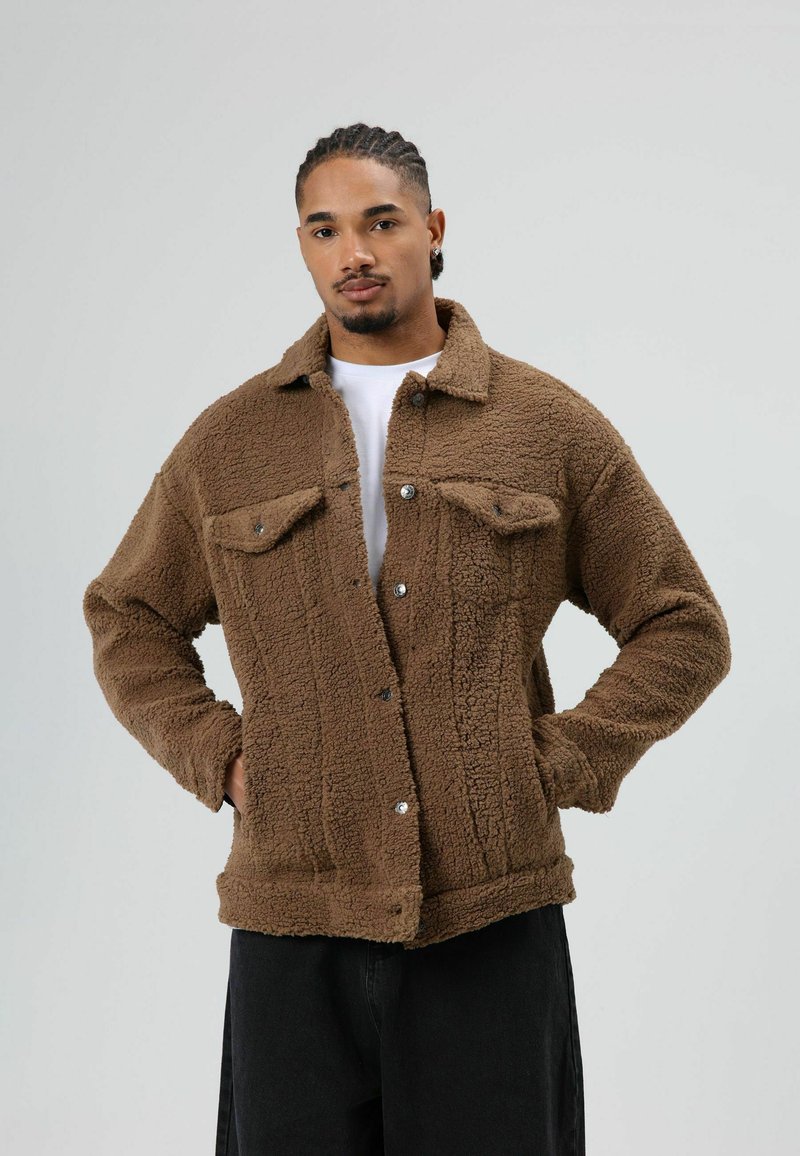 Brown fleece jacket with a textured surface, button closure, and two chest pockets. Model wears a white shirt and black pants.