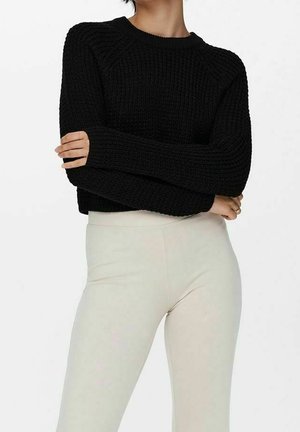 Schwarzer, gestrickter Pullover mit Rundhalsausschnitt und geripptem Muster, mit langen Ärmeln und einem kurzen Schnitt, kombiniert mit hellfarbenen, enganliegenden Hosen.