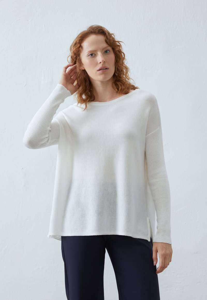 Pulover alb cu mâneci lungi asemănătoare cu un model încrețit și slituri pe laterale. Textură moale și croială lejeră, asortat cu pantaloni închisi la culoare. Design minimalist.