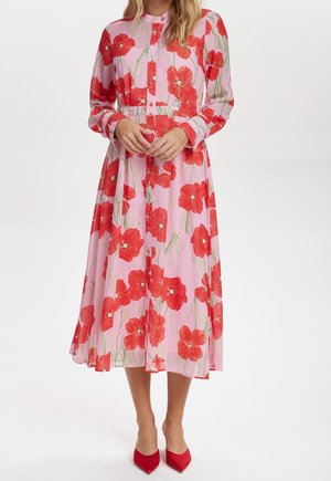 Femme portant une robe midi rose à manches longues avec un motif floral rouge et des talons rouges à bouts pointus sur fond blanc.