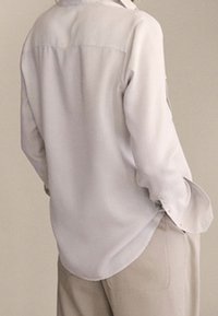 Jasnoszara bluzka z długim rękawem wykonana z gładkiego materiału, charakteryzująca się prostym krojem, guzikami przy mankietach oraz zaokrąglonym dołem.