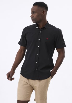 Camisa negra de manga corta con cuello abotonado, que cuenta con botones blancos y un pequeño logo rojo en el pecho. Se lleva con pantalones cortos beige.