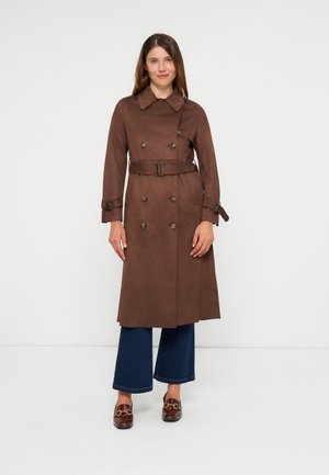 Femme portant un long manteau marron croisé avec ceinture, un jean bleu foncé à jambes larges et des mocassins marron sur un fond blanc uni.
