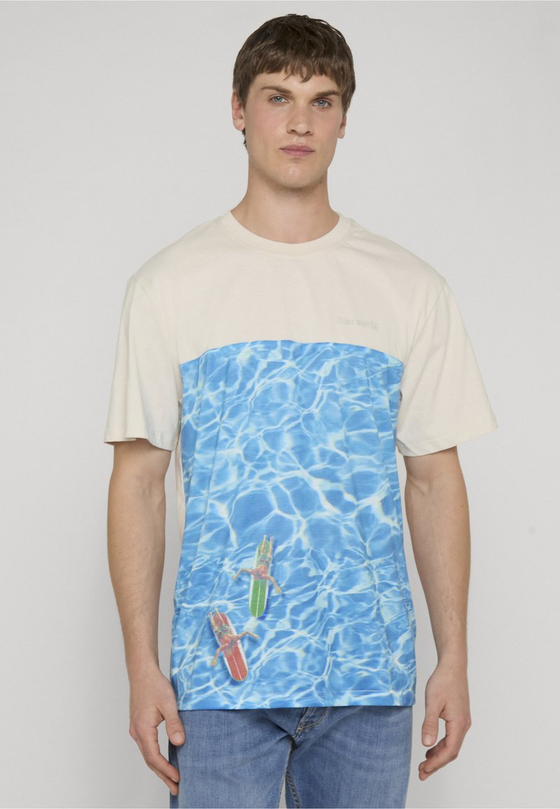 Homme portant un T-shirt beige et bleu avec un graphisme d'eau et deux surfeurs, associé à un jean bleu, debout devant un fond uni.