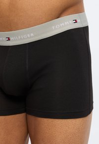 Μαύρα εφαρμοστά αντρικά μποξέρ με γκρι ελαστική μέση που φέρει το λογότυπο "Tommy Hilfiger", φορεμένα στη χαμηλή κοιλιά και τα πάνω μηρά ενός ατόμου.