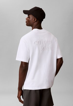 Junger Mann trägt ein weißes Calvin Klein T-Shirt und eine schwarze Kappe, seitlich vor einem schlichten grauen Hintergrund.