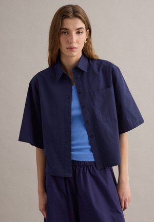 Giovane donna con capelli lunghi indossa una camicia blu navy a maniche corte con bottoni, ampia, sopra una canottiera blu aderente e pantaloni blu navy abbinati.