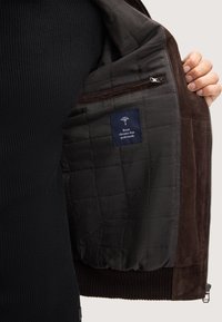 Binnenzijde van een bruine suède jacket met een gewatteerde voering, een ritszak en een stoffen label. Geribbelde randen en een gladde textuur verhogen de warmte en het comfort.