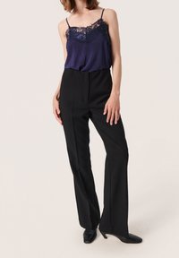 Camisola di raso blu navy con pizzo, abbinata a pantaloni neri a vita alta con gamba affusolata e texture liscia, indossata con tacchi neri.