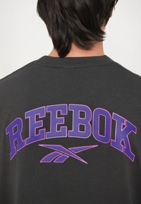 Camiseta de algodón gris con un gran logo púrpura de "REEBOK" en la parte posterior, con acentos en rosa y un diseño de cuello redondo.