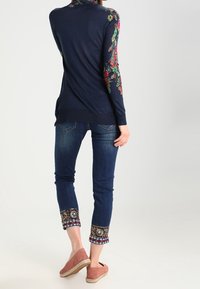Marineblaues Langarmshirt mit Blumenstickerei an den Ärmeln, kombiniert mit dunklen Jeans mit bunten, gemusterten Bündchen und hellbraunen Espadrilles.