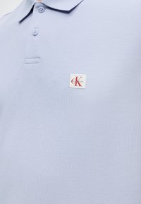 Φωτεινό γαλάζιο πόλο με δύο κουμπιά και ένα μικρό λευκό ορθογώνιο έμβλημα με κόκκινα και γκρι γράμματα στο στήθος.