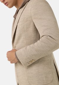 Béžový blazer s texturovanou tkaninou, s dvěma knoflíky a náprsní kapsou; design s kapucí a kontrastní hnědou podšívkou.