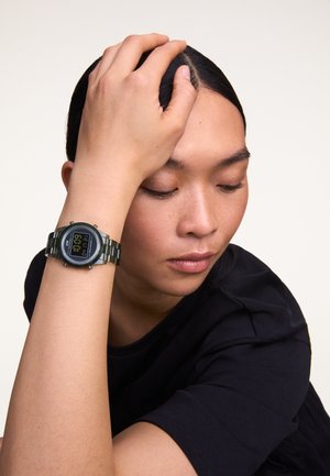 Reloj digital con esfera negra y pantalla blanca, con una pulsera de metal en verde oscuro, llevado en un brazo con una textura de piel suave.